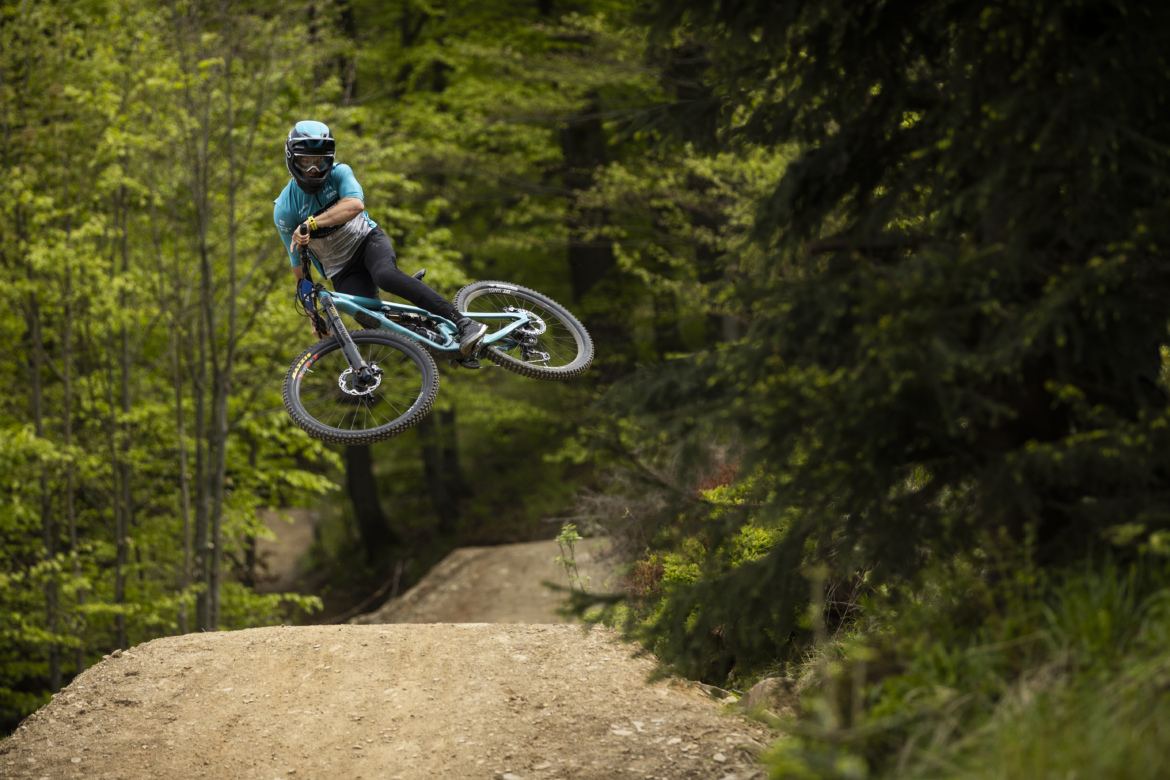 A person wearing a helmet and blue and black riding gear performing a dirt bike jump in a forested area with lush green trees in the background.