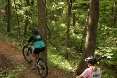 Two cyclists navigate a dirt trail through a lush, green forest. The first rider, wearing a teal and black shirt, is positioned in the foreground, while the second, dressed in a pink shirt, follows behind. Tall trees surround them, creating a serene and natural setting. Sunlight filters through the leaves, highlighting the vibrant greenery of the woodland area. Bobcat Trail mountain bike trail.