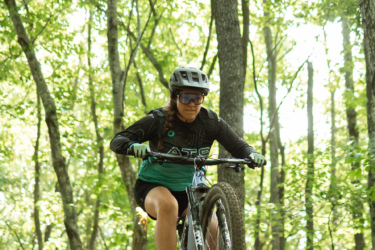 A young woman wearing a helmet and sunglasses rides a mountain bike over a dirt trail in a lush green forest. She is performing a jump, with the front wheel of the bike lifted off the ground, surrounded by tall trees and greenery in the background. The sunlight filters through the leaves, creating a vibrant and dynamic atmosphere. Bobcat Trail mountain bike trail.