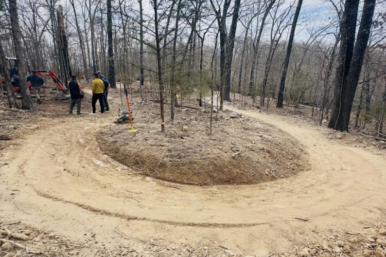 Two people work on a newly constructed trail in a wooded area, featuring a sandy, curved path that leads around a raised dirt mound. Leafless trees surround the scene under a cloudy sky.