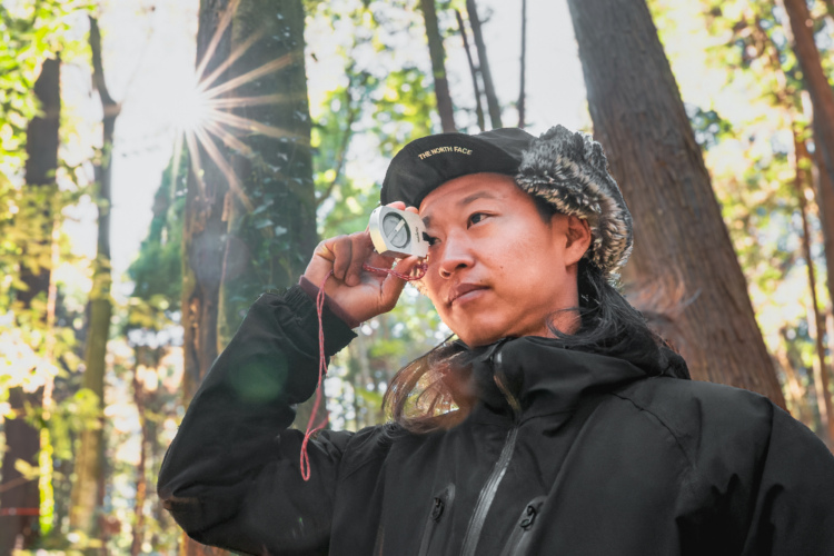 A person wearing a black jacket and a hat stands in a forest, holding a compass to their forehead while looking thoughtfully into the distance. Sunlight filters through the trees, creating a natural lens flare in the image.