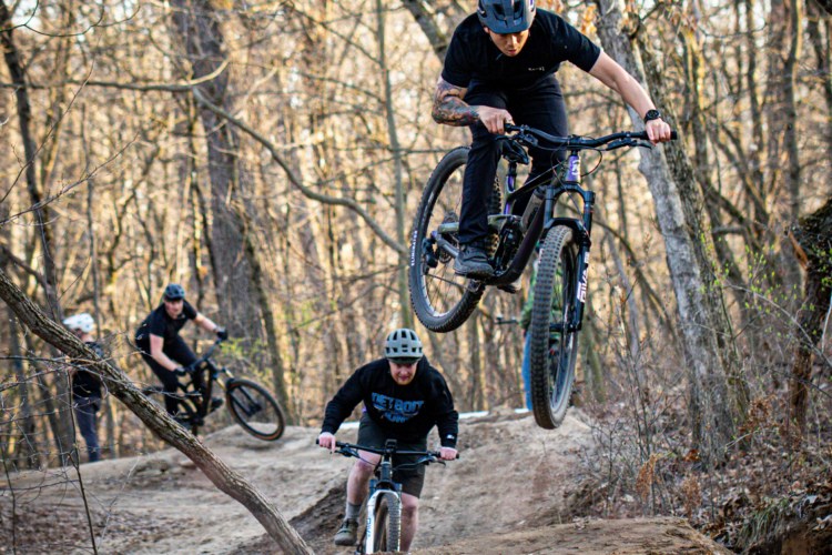 Three individuals are riding mountain bikes on a dirt trail in a forested area. The main rider is airborne, performing a jump, while two others navigate the trail behind. The scene captures the excitement of mountain biking amid trees with bare branches, indicating an early spring or late fall setting.