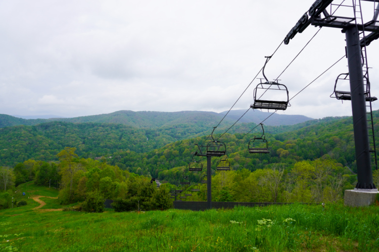 A scenic view of a lush green landscape featuring a ski lift in the foreground. The lift chairs are empty, ascending toward soft, rolling hills covered in vibrant green trees under a cloudy sky. The image captures the tranquility of nature in a mountainous area.