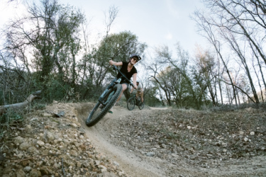 Two mountain bikers navigate a dirt trail surrounded by trees. One rider, wearing a helmet and a black shirt, is leaning into a turn with a big smile, while the second rider follows behind on the path. The scene captures a sense of adventure and enjoyment in a natural outdoor setting. Swasey Recreation Area mountain bike trail.