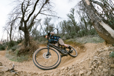 A mountain biker leans into a dirt turn on a rugged trail surrounded by trees. The biker is wearing a colorful shirt and protective gear, showcasing an energetic riding style. The scene captures the essence of outdoor adventure and the thrill of mountain biking. Swasey Recreation Area mountain bike trail.