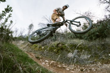 A mountain biker in a helmet jumps over a dirt trail, capturing air while riding on a rocky path surrounded by greenery and sparse trees. Swasey Recreation Area mountain bike trail.