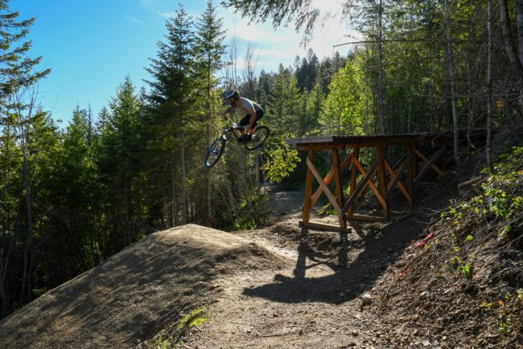 A cyclist performs a jump on a bike over a dirt ramp, surrounded by lush green trees and a clear blue sky. A wooden structure is visible nearby, providing a landing spot for the jump.