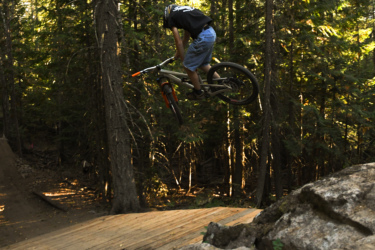 A mountain biker performs a jump off a wooden ramp in a forested area, surrounded by tall trees. The rider, wearing a black t-shirt and denim shorts, is mid-air, showcasing their skill in a dynamic pose. Sunlight filters through the trees, creating a vibrant natural backdrop. Missing Lynx mountain bike trail.