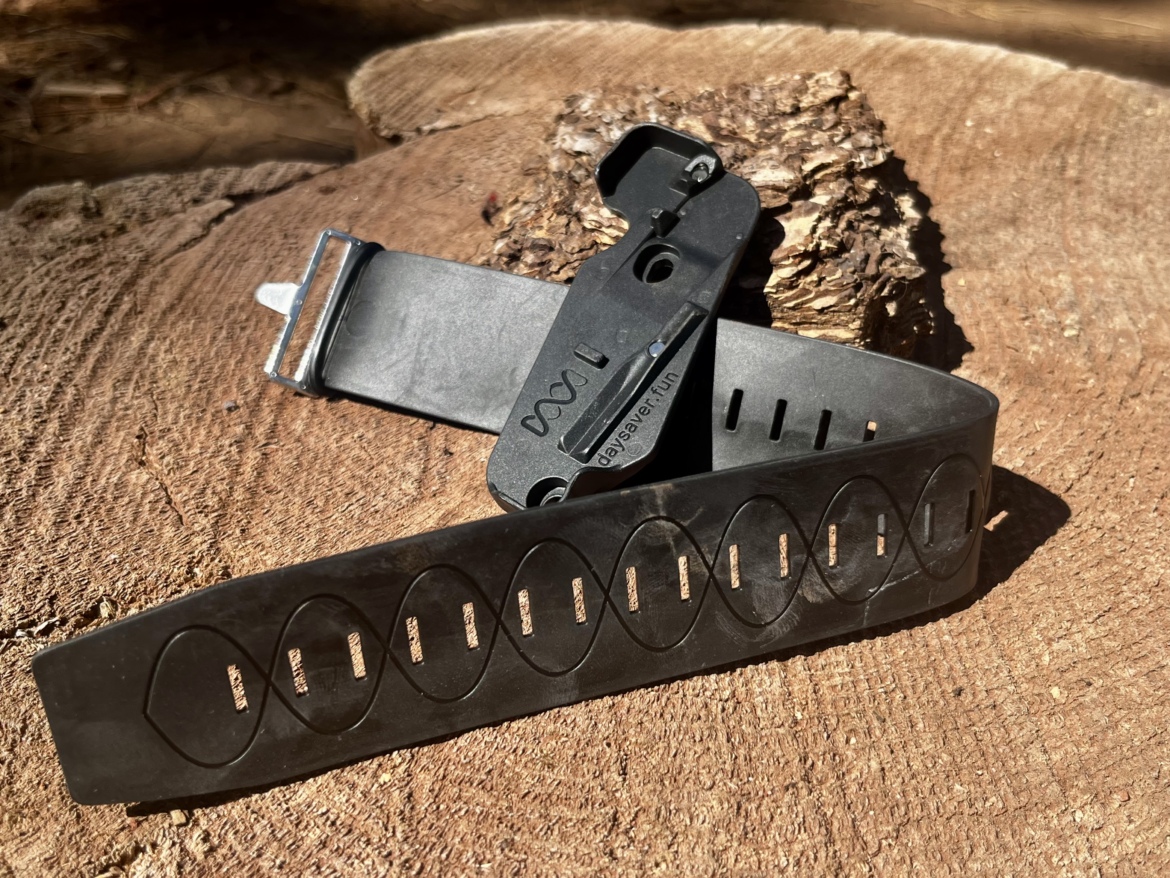 A close-up image of a black rubber wristband with a unique wavy pattern and a small black clamshell-style buckle resting on a wooden surface. The background features a rustic wooden log, enhancing the natural setting of the scene.