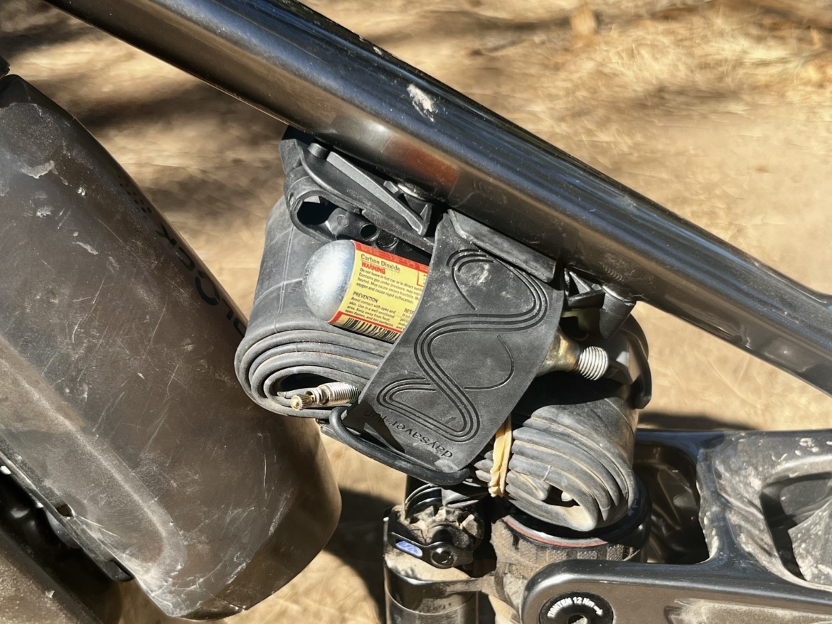 Close-up of a bicycle suspension system, featuring a black air compressor with rubber hoses and a label. The image shows the compressor mounted securely to the bike frame, with dirt visible on the frame indicating outdoor use. The background is a blurred earth-tone landscape.