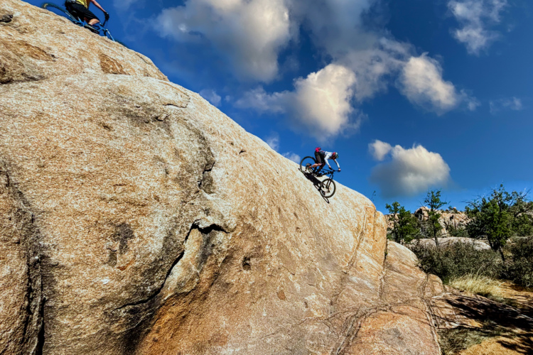 Two mountain bikers navigate steep rocky terrain against a backdrop of blue sky and fluffy clouds. One biker descends a large granite rock, while the other ascends from a higher vantage point. Vegetation and additional rock formations are visible in the surrounding landscape.