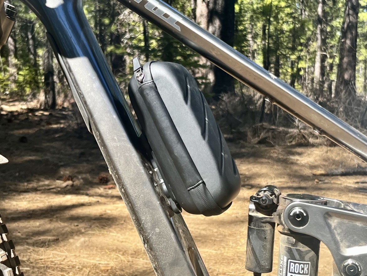 Close-up image of a bike frame with a black storage box attached to the side. The background features a forest setting with trees and pine needles on the ground.