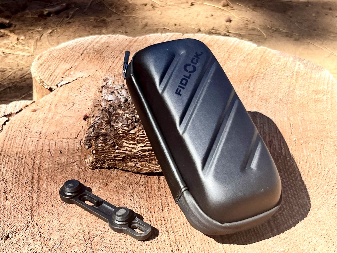 Black hard-shell case with a textured surface, featuring the brand name "FIDLOCK," resting on a wooden log in an outdoor setting. A small black attachment piece is placed nearby.
