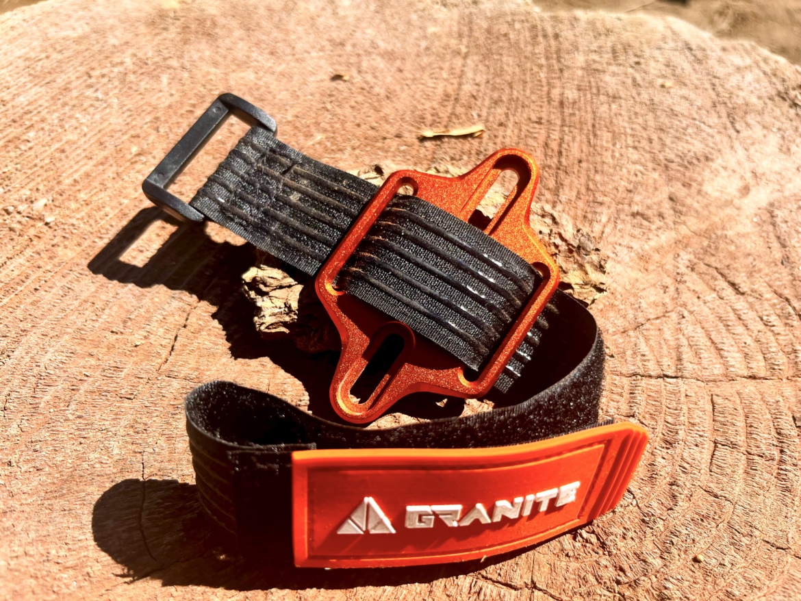 A close-up image of a black and orange strap with a metal buckle, resting on a wooden surface. The strap features a logo that reads "GRANITE" and has a textured black design with an orange accent. The lighting highlights the details of the materials and the natural wood background.