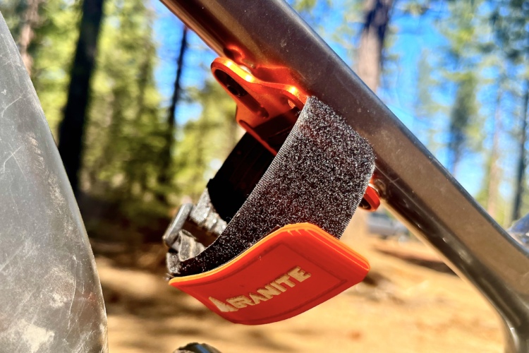 Close-up image of a metallic bicycle frame featuring a bright orange clip attached to it. The clip has a black padded section and the word "LARGANITE" is visible on the orange part. In the background, there are trees suggesting an outdoor setting.