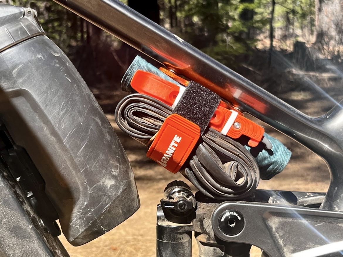 Close-up of a bike frame showing a mounted roll of cables secured with orange straps and a black Velcro strip. A section of the bike's rear suspension is visible, along with part of the tire and the surrounding forest backdrop.