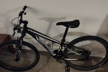 Specialized Hardrock 26: A black and white mountain bike is leaning against a wall on a carpeted floor. The bike features thick tires, a streamlined frame, and standard handlebars. Nearby, there is a wooden piece of furniture partially visible in the image.