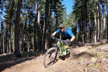 A mountain biker in a blue shirt and helmet skillfully navigates a winding dirt trail in a forested area, surrounded by tall trees and a clear blue sky. Dust is kicked up from the ground as he leans into the turn on his green bike. Mount Ashland Shuttle mountain bike trail.