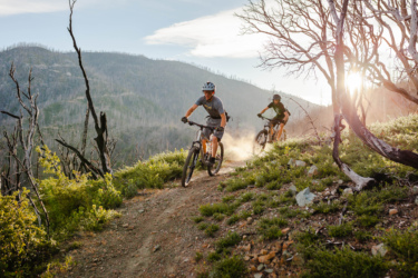 Two mountain bikers riding along a dusty trail through a lush, green landscape, with a backdrop of mountains and trees. The sun is setting, casting a warm glow over the scene, and there are signs of burnt trees in the area. Swasey Recreation Area mountain bike trail.