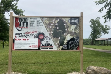 A wooden sign at Quarry Park displaying a map of Wicked Walt's Mountain Bike Trail. The sign includes trail details, safety information for riders, and is surrounded by grassy areas and trees. A picnic shelter is visible in the background. Wicked Walt’s mountain bike trail.