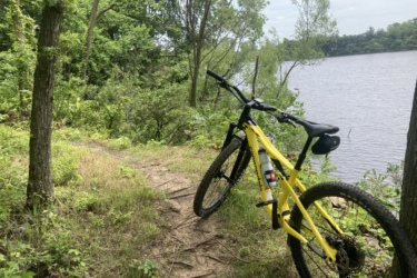 A yellow mountain bike is parked next to a small trail leading to a calm lake, surrounded by lush green vegetation and trees. The sky is partially cloudy, suggesting a pleasant day for outdoor activities. Wicked Walt’s mountain bike trail.