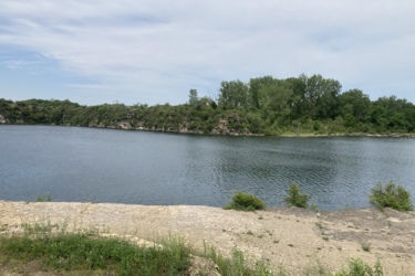 A serene view of a calm lake bordered by lush greenery and rock formations under a partly cloudy sky. The still water reflects the surrounding trees and cliffs, creating a peaceful natural landscape. Wicked Walt’s mountain bike trail.