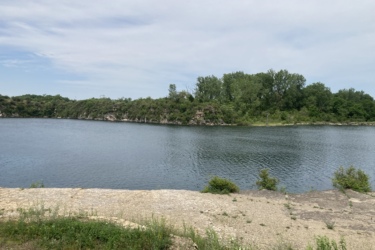 A tranquil lakeside scene featuring calm water surrounded by lush greenery and rocky cliffs under a partly cloudy sky. The foreground includes a rocky shoreline with patches of grass and small plants. Wicked Walt’s mountain bike trail.