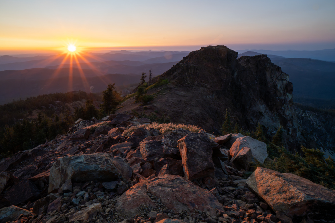 A breathtaking sunrise over a mountainous landscape, with the sun's rays illuminating the rocky foreground and distant hills. Evergreen trees dot the scene, adding to the natural beauty of the tranquil setting.