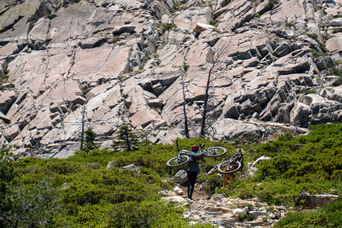 A person wearing a helmet and carrying a bicycle walks along a rocky trail surrounded by lush greenery and steep rock formations. Another bicycle is being carried by a companion nearby. Sunlight illuminates the scene, creating a vibrant outdoor atmosphere.