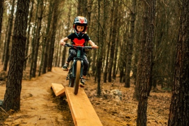 A young child in a helmet rides a bike along a wooden plank elevated above a dirt trail, surrounded by tall trees in a forest setting. The child appears focused and balanced as they navigate the narrow path. Lakewoods Trail System mountain bike trail.