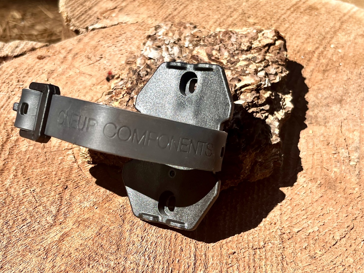 Black bike component mounted on a textured wooden surface, featuring the text "ONEUP COMPONENTS". The component has a rectangular shape with a strap attached, designed for securing to a bicycle frame or accessory.