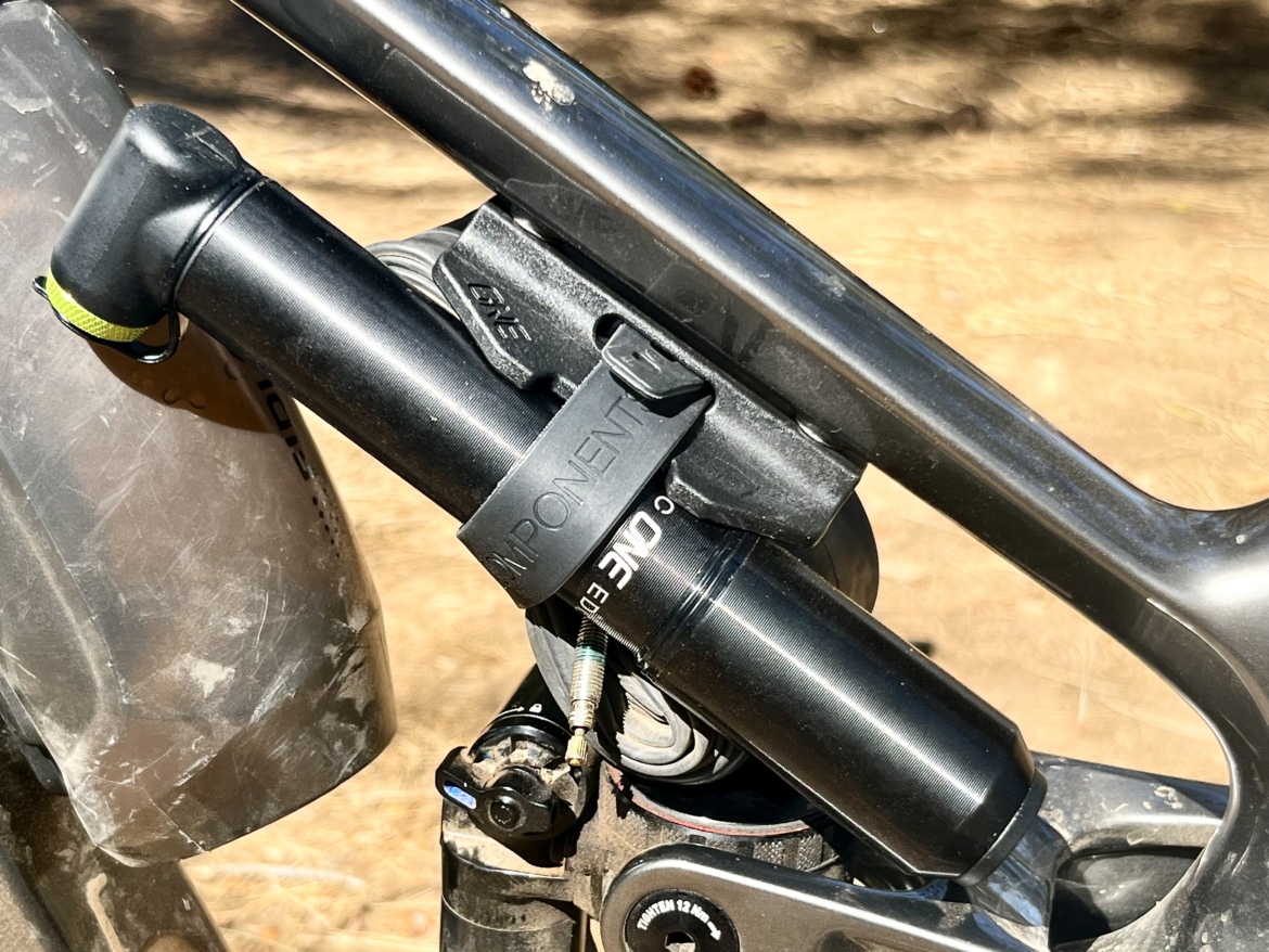Close-up of a mountain bike shock absorber mounted on the frame, featuring a black cylindrical body with a label and a yellow securing strap, against a background of a dusty terrain.