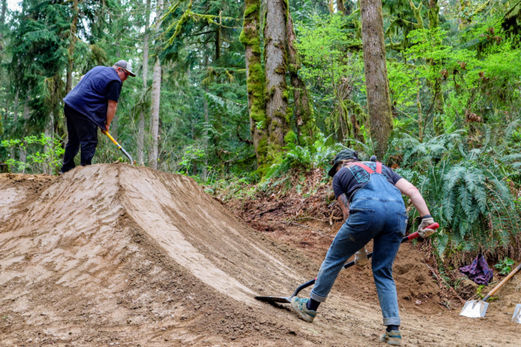 Two people working on building a dirt ramp in a forested area. One person is using a shovel and is standing on top of the ramp, while the other is kneeling and shaping the dirt with a larger shovel. Surrounding them are tall trees and greenery, creating a natural outdoor setting.