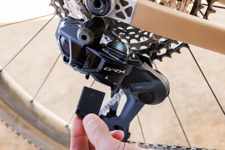 A close-up image of a bicycle's rear derailleur, featuring a Shimano GRX component. A hand is seen holding a small black piece, likely a battery or connector, that is being prepared for installation or adjustment, with the bicycle chain and cassette in the background.
