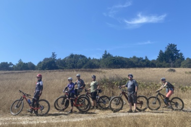 A group of six cyclists poses on mountain bikes along a gravel path surrounded by tall, golden grass and trees under a clear blue sky.