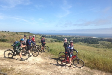 A group of seven mountain bikers paused on a dirt trail, surrounded by lush green hills and a view of the ocean in the background. The cyclists, wearing helmets and various biking gear, are positioned on their bikes, enjoying the scenic landscape under a partly cloudy blue sky.