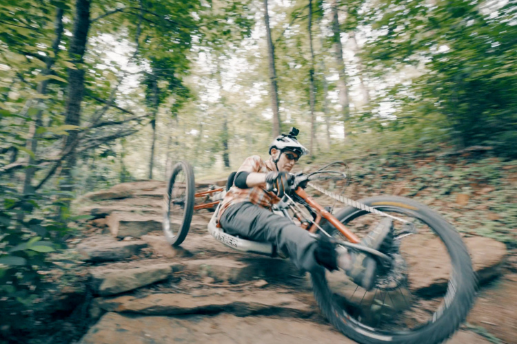 A cyclist navigating rough terrain on a mountain bike, leaning to one side as they traverse a rocky path in a wooded area. The image conveys a sense of speed and movement, with trees and foliage blurred in the background.