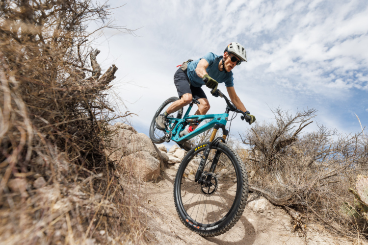 A mountain biker navigating a rocky trail, maneuvering down a slope with focus and determination. The rider is wearing a helmet and sunglasses, and the bike features a teal frame and knobby tires, set against a backdrop of dry, rugged terrain and a cloudy sky.