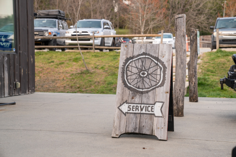 A wooden A-frame sign displaying a bicycle wheel graphic and the word "SERVICE," pointing left. In the background, several vehicles are parked, with a grassy area and trees visible.