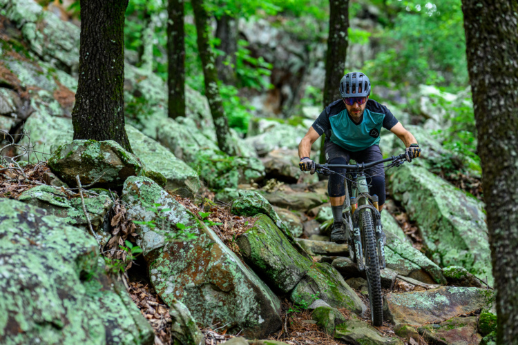 A mountain biker navigating a rocky trail in a lush green forest, wearing a helmet and protective gear, focusing on maintaining balance while riding over uneven terrain.