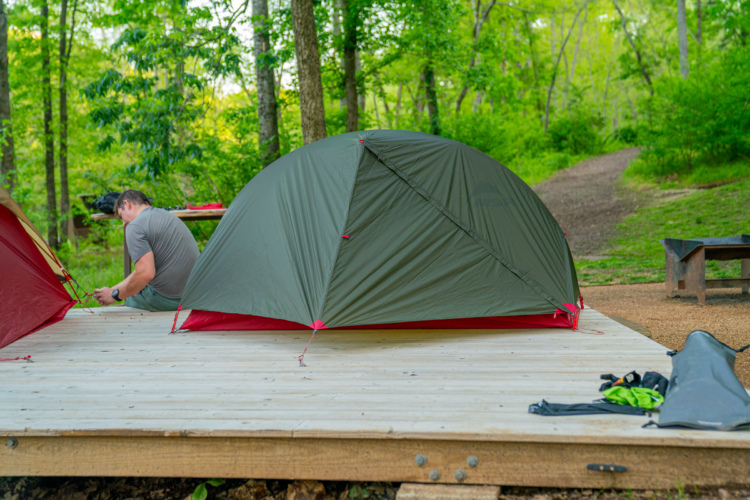 A campsite featuring a green and red tent set up on a wooden platform, with a person sitting nearby, engaged in outdoor activity. Lush trees and foliage provide a natural backdrop, with a pathway visible in the background. Camping gear is scattered around the scene.