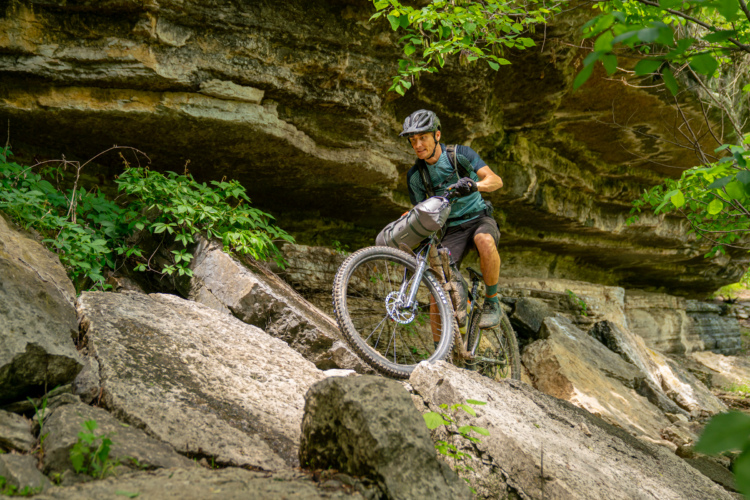 A mountain biker navigates over rocky terrain beneath a rocky overhang, surrounded by lush greenery. The rider is wearing a helmet and a backpack, focusing intently on the trail ahead.