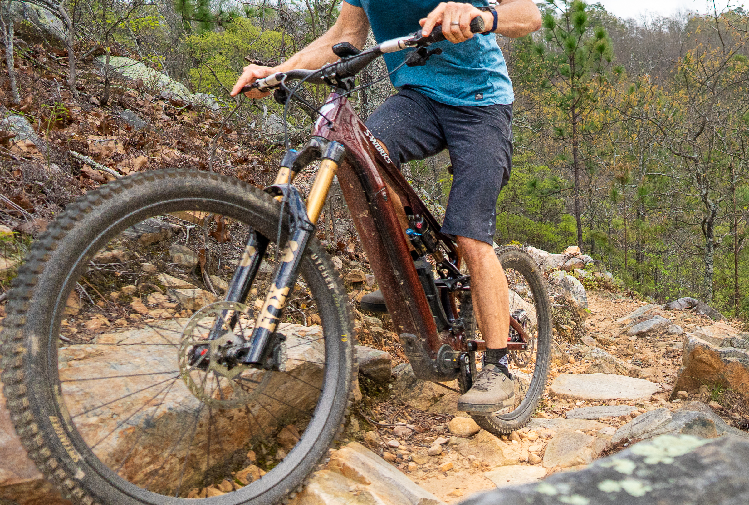 A cyclist rides an electric mountain bike on a rocky trail surrounded by trees and greenery. The biker is wearing a blue shirt and black shorts, showcasing an intense focus as they navigate the rugged terrain. The scene captures the spirit of outdoor adventure and mountain biking.