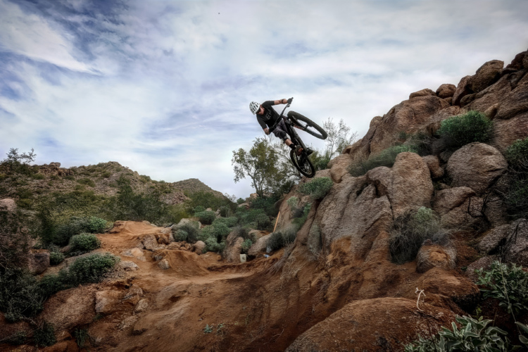 A mountain biker performing a jump over rocky terrain, surrounded by desert vegetation and hills under a cloudy sky.