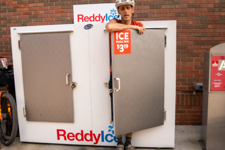 A person in a helmet leans out of a Reddy Ice ice vending machine, with an open door displaying a price sign for small bags of ice at $3.19. A bike is partially visible in the background, and the machine is set against a brick wall.