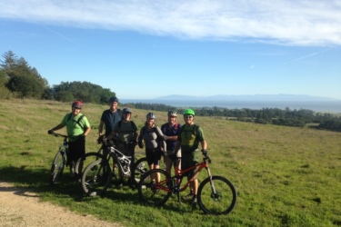 A group of six mountain bikers poses for a photo on a grassy hilltop with a scenic view of rolling hills and the ocean in the background. The cyclists are wearing helmets and riding gear, with their bikes beside them. The sky is clear with a few clouds, indicating a bright and sunny day.