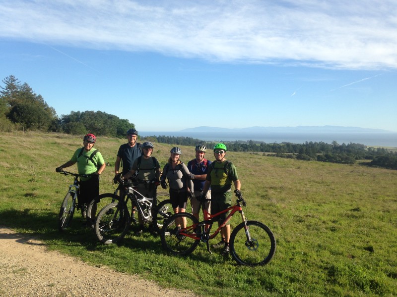 A group of six mountain bikers poses for a photo on a grassy hilltop with a scenic view of rolling hills and the ocean in the background. The cyclists are wearing helmets and riding gear, with their bikes beside them. The sky is clear with a few clouds, indicating a bright and sunny day.