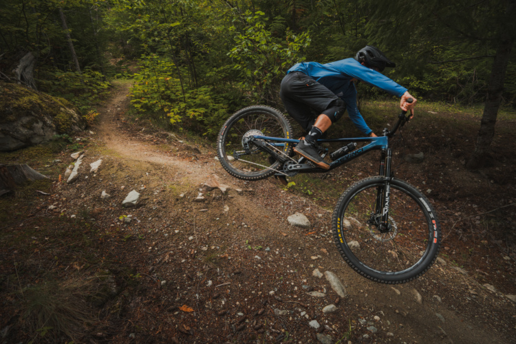 A mountain biker in blue gear leans into a bend on a dirt trail surrounded by lush greenery, showcasing an action-packed moment of cycling on rugged terrain.