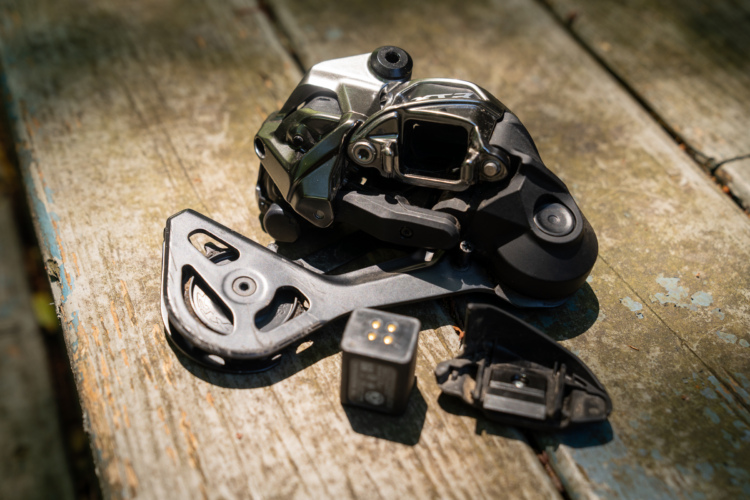 A close-up image of a bicycle derailleur assembly, including the main derailleur body, a lower pulley, and a small electronic component, all placed on a weathered wooden surface. The derailleur features a combination of metallic and black plastic parts, emphasizing its mechanical design.