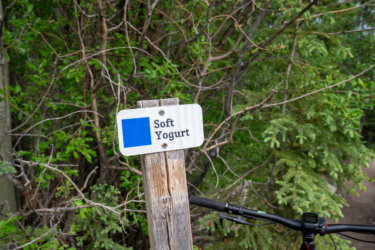 A wooden signpost in a natural setting, featuring the text "Soft Yogurt" and a blue square. In the foreground, part of a bicycle handlebar is visible, surrounded by greenery and trees. Soft Yogurt mountain bike trail.